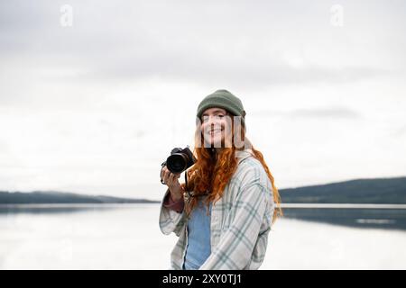 Una gioiosa donna dai capelli rossi che indossa un comodo cappello tiene in mano una macchina fotografica, pronta a catturare momenti in un lago sereno sotto un cielo nuvoloso, raffigurando perfettamente la pace Foto Stock