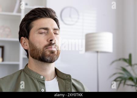 Uomo barbuto a occhi chiusi che gode di momenti di relax e tranquillità, concentrandosi sulla consapevolezza in un ambiente tranquillo e familiare. Sollievo dallo stress e stato mentale pacifico. Foto Stock