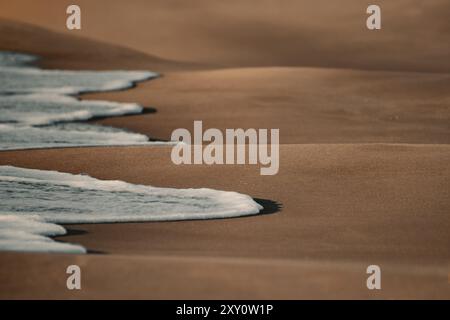 Una scena tranquilla che cattura la delicata schiuma delle onde marine mentre incontrano delicatamente la sabbia liscia di una spiaggia tranquilla, mettendo in risalto le texture della natura. Foto Stock