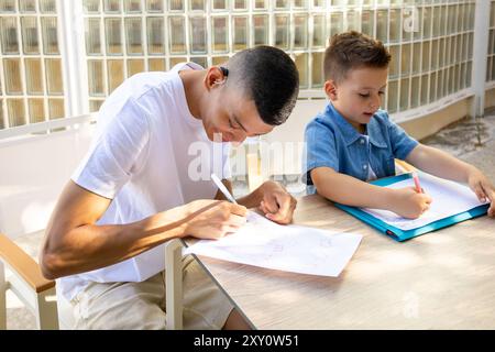 Un giovane uomo con paralisi cerebrale e sordità lieve impegnato a disegnare con un ragazzo a un tavolo illuminato dal sole, mostrando inclusione e creatività Foto Stock