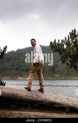 Un giovane sorride mentre salta su un tronco caduto vicino a un lago nebbioso, circondato da una lussureggiante pineta, esplorando la bellezza naturale di un giorno grigio e piovoso Foto Stock