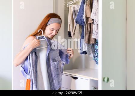 Una giovane donna sta organizzando il suo guardaroba, esaminando attentamente i vestiti da mettere in vendita. Sta guardando in basso, è pienamente impegnata nel suo compito. Foto Stock