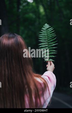 Vista posteriore di una donna irriconoscibile in una giacca rosa che interagisce con la natura mentre tiene con cura una foglia di felce, circondata da una fitta foresta verde. Foto Stock