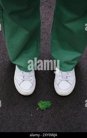 Vista corta e irriconoscibile dei piedi di una persona con sneakers bianche e pantaloni verdi in piedi su un sentiero scuro con una distinta foglia verde sulla groun Foto Stock