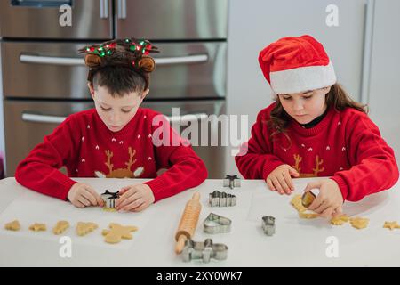 Due bambini, vestiti con maglioni rossi e cappelli natalizi, si concentrano intensamente sul taglio di biscotti a forma di festa in una cucina moderna Foto Stock