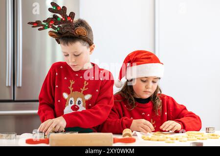 Gemelli in maglioni per le vacanze e cappelli di Babbo Natale che preparano biscotti in cucina, concentrandosi intensamente sulla forma dell'impasto Foto Stock