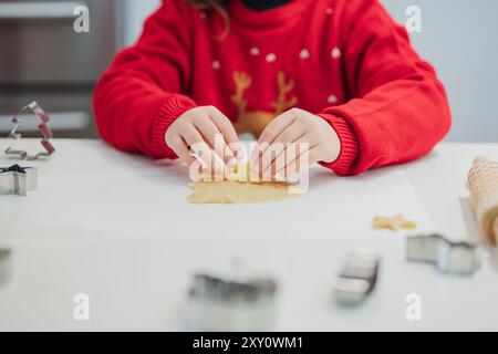 Un bambino in un maglione rosso vivace prepara i biscotti di Natale in un ambiente accogliente, utilizzando una varietà di forbici sull'impasto Foto Stock