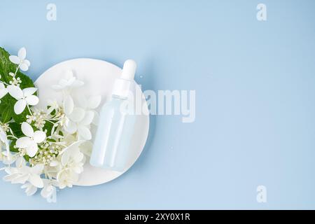 Vista dall'alto di una serena bottiglia di siero per la cura della pelle accanto a delicati fiori bianchi e un piatto in ceramica su un tranquillo sfondo blu che cattura l'essenza del pu Foto Stock