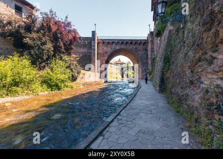 Vista posteriore di una persona irriconoscibile che cammina sotto un ponte di pietra sul fiume nell'affascinante villaggio di Potes, con vegetazione lussureggiante e traditiona Foto Stock