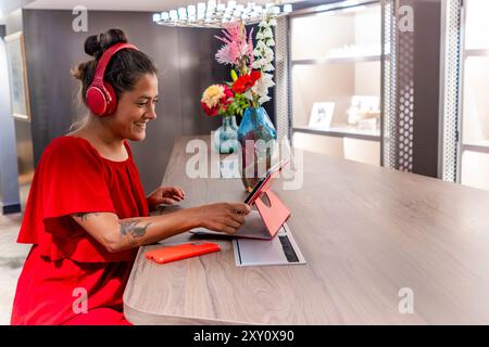 Una donna gioiosa vestita con un vivace abito rosso ama lavorare in una moderna scrivania da ufficio, dotata di un computer portatile, cuffie e smartphone Foto Stock