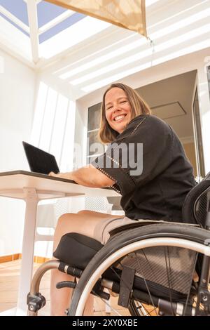 Un'allegra donna d'affari in sedia a rotelle lavora sul suo notebook da una casa luminosa, dimostrando produttività e determinazione Foto Stock