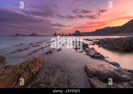 Una tranquilla alba bagna Gueirua Beach, le Asturie, in tonalità calde, mentre le formazioni rocciose frastagliate e le acque calme catturano l'essenza della bellezza costiera. Foto Stock