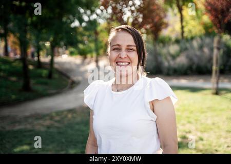 Una gioiosa donna colombiana con camicetta bianca si erge in un parco illuminato dal sole, sorridendo calorosamente alla macchina fotografica. Lo sfondo mostra un percorso sfocato e un ambiente lussureggiante Foto Stock