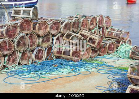 Una pila di trappole da pesca cilindriche, alcune racchiuse in reti, accanto a gruppi intrecciati di corde blu e verdi su un molo di cemento, con una barca e un wat Foto Stock