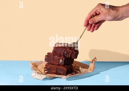 Persona corta e irriconoscibile che versa una ricca salsa di cioccolato su una torre di brownie al cioccolato fatti in casa, serviti su carta pergamena con un dorso bicolore Foto Stock
