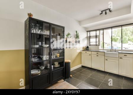 Una cucina moderna ben illuminata con un classico armadio nero pieno di piatti e bicchieri, armadi color crema e una finestra con persiane. Foto Stock