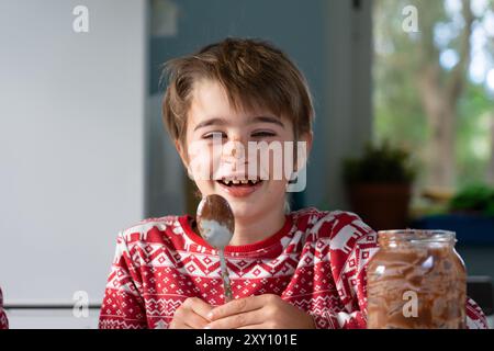 Ritratto di un ragazzo caucasico ridendo con la faccia spalmata di cioccolato mentre preparava il dessert natalizio vestito con abiti natalizi Foto Stock
