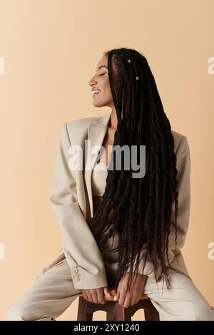 Un'elegante donna afroamericana siede su uno sgabello, indossa un abito beige e sorride. Foto Stock