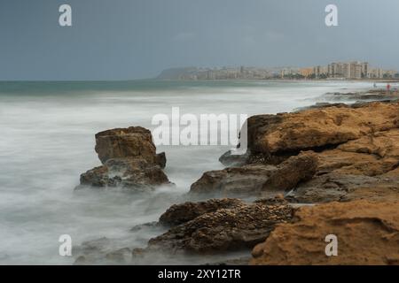 Fotografie a lunga esposizione sulla costa mediterranea con un mare tempestoso e sullo sfondo Urbanova e Arenales del Sol, Alicante, Spagna Foto Stock