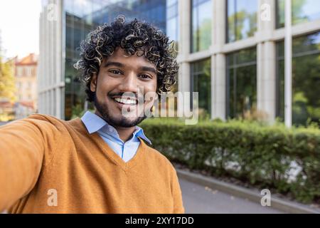 Uomo con capelli ricci che scatta selfie in città. Indossa un maglione arancione sopra una camicia blu. Sorridendo in senso lato, godendo dell'ambiente all'aperto. La tecnologia enfatizza la natura amichevole e accessibile. Foto Stock