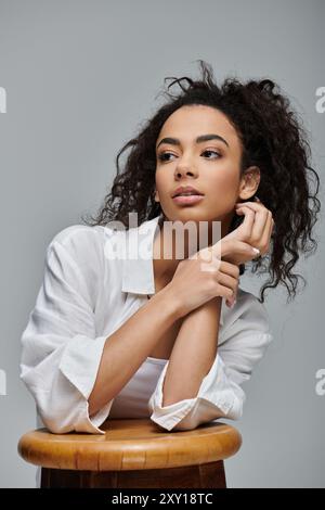 Una giovane donna afroamericana con i capelli ricci si siede su uno sgabello di legno su uno sfondo grigio, vestito con una camicia bianca con colletto. Foto Stock