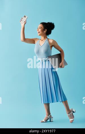 Una giovane donna con un vestito blu sorride brillantemente mentre tiene in mano un laptop e un telefono in un ambiente studio. Foto Stock