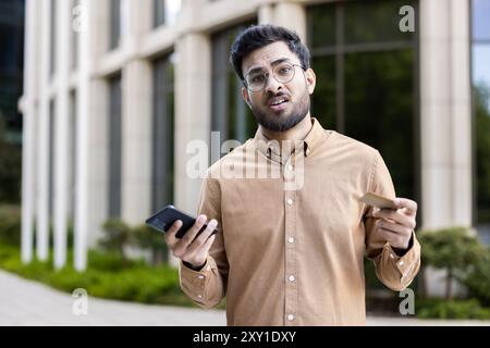 Uomo con occhiali in piedi fuori, con telefono e carta di credito, che mostra espressioni confuse. Concetto di incertezza finanziaria, frustrazione tecnologica, processo decisionale o problemi di comunicazione. Foto Stock
