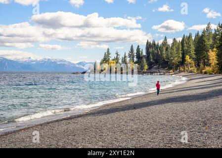 Donna solitaria in giacca con cappuccio rossa che cammina su una spiaggia di ciottoli su un lago in una fredda giornata autunnale. Foto Stock