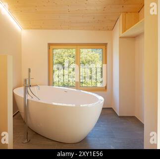 Vista frontale di una moderna vasca da bagno in un bagno con soffitto in legno e una luminosa finestra affacciata sulla natura. Nessuno dentro Foto Stock