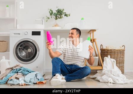 Giovane felice con bottiglie di detergente e pile di vestiti sporchi seduti sul pavimento nella lavanderia Foto Stock