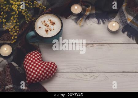 Cappuccino caldo, coperta, candele, fiori di mimosa e cuore di tessuto su vecchio sfondo in legno. Tonning vintage. Concetto di relax romantico stagionale, fl Foto Stock