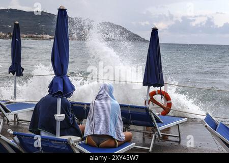 Onde che si infrangono contro il molo di un molo dove una coppia è seduta su sdraio, Diana Marina, Italia, 18/08/2024, Diano Marina, Liguria, IT Foto Stock