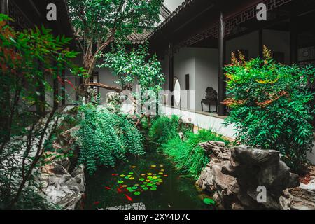 Un lussureggiante giardino verde con un piccolo laghetto circondato da rocce, alberi e tradizionale architettura cinese. Foto Stock