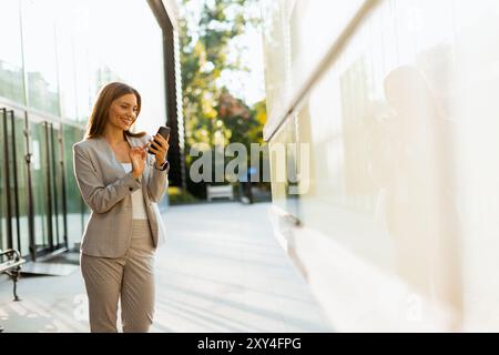 Una donna professionista cammina con facilità, sorridendo mentre si concentra sul telefono, mescolando lo stile di vita moderno con la tranquillità della natura sullo sfondo Foto Stock