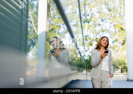 Una donna professionista cammina con facilità, sorridendo mentre si concentra sul telefono, mescolando lo stile di vita moderno con la tranquillità della natura sullo sfondo Foto Stock