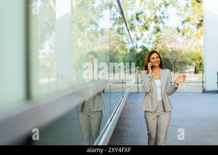 Una donna professionista cammina con facilità, sorridendo mentre si concentra sul telefono, mescolando lo stile di vita moderno con la tranquillità della natura sullo sfondo Foto Stock