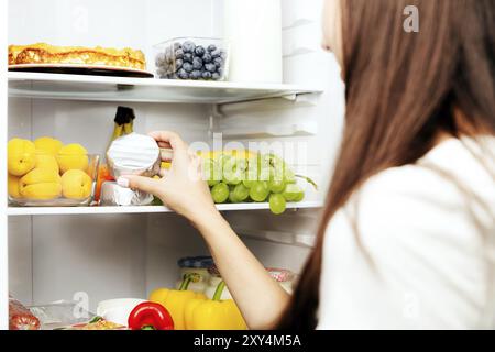 Donna che prende a mano, afferra o preleva il camembert o il formaggio brie dal ripiano del frigorifero aperto, un cassetto del frigorifero pieno di frutta, verdura, banana, piselli Foto Stock