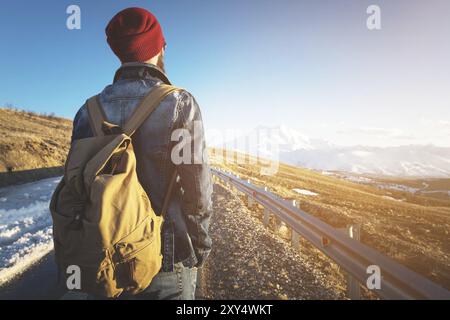 Un hipster barbuto con uno zaino vintage vecchio stile che indossa occhiali da sole con un cappello rosso e una giacca di jeans e jeans si erge su un asfalto di campagna Foto Stock