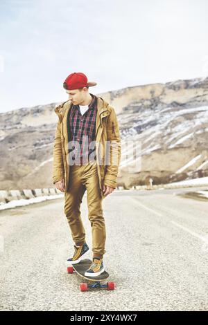 Elegante giovane felice con un berretto e pantaloni che jogger girovagano lungo una strada di montagna su un longboard, godendosi la vita Foto Stock