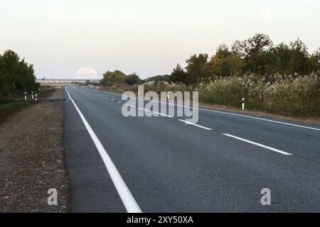 Un'enorme luna piena al mattino presto che lascia l'orizzonte sopra la strada asfaltata. Foto vera Foto Stock