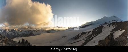 Un panorama al tramonto dell'elbrus e parte della cresta caucasica con nuvole arancioni e un ghiacciaio incrinato sul fondo Foto Stock