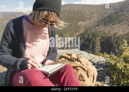 Ritratto di una ragazza hipster che indossa occhiali da sole e un cappello seduto su una roccia all'aperto in montagna contro un cielo blu. Designer freelance in viaggio d Foto Stock