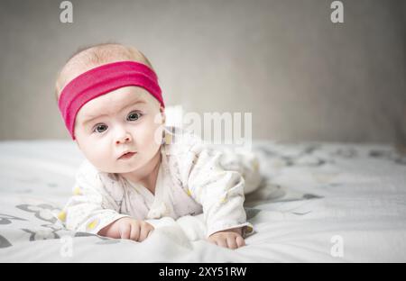 Adorabile bambina di 6 mesi sul letto sulla pancia con la testa in alto che guarda la telecamera con i suoi grandi occhi. Luce naturale della camera da letto Foto Stock