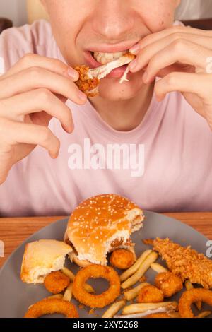 Uomo caucasico che mangia ali di pollo fritte dorate, hamburger, patatine fritte, anelli di cipolla e pezzi di ali di pollo in piatto grigio su un tavolo di legno, americano Foto Stock