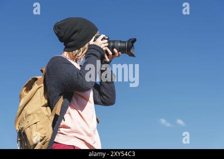 Ritratto di una ragazza hippy elegante con un cappello e uno zaino che la fotografa con una fotocamera reflex digitale all'aperto contro un cielo blu in una zona soleggiata Foto Stock