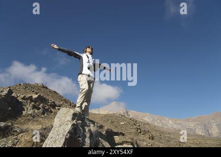 Un applauso di una giovane donna in spalla sulla cima di un'ampia cima di montagna. Libertà e vittoria sullo sfondo di montagne e blu Foto Stock