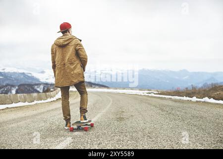 Elegante giovane felice con un berretto e pantaloni che jogger girovagano lungo una strada di montagna su un longboard, godendosi la vita Foto Stock