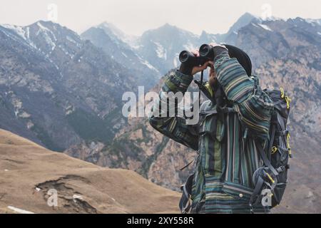 Un uomo hipster con barba in cappello, giacca e zaino nelle montagne caucasiche contiene binocoli, avventura, turismo, tracciamento Foto Stock