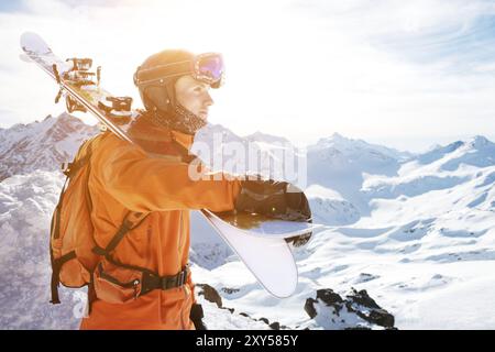 Ritratto di uno sciatore in una tuta arancione con uno zaino sulla schiena e gli sci sulle spalle in un casco si erge su una roccia contro il cielo blu. Copia Foto Stock