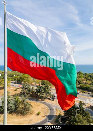 La bandiera bulgara ondeggia orgogliosamente sopra una città costiera, con il vasto mare e il verde paesaggio sullo sfondo. Un simbolo di orgoglio nazionale e.. Foto Stock